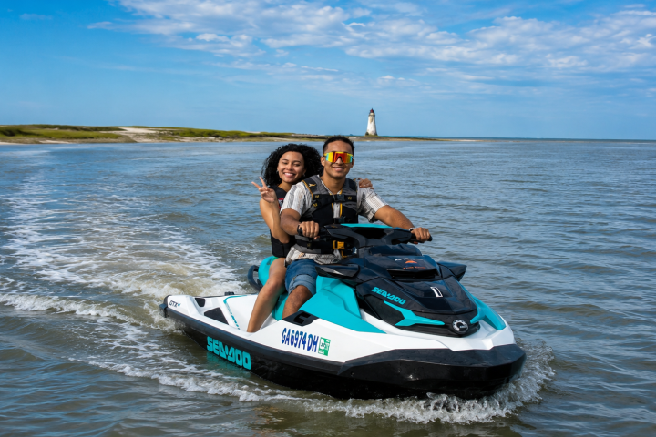 Two people riding a jet ski on open water near a lighthouse.