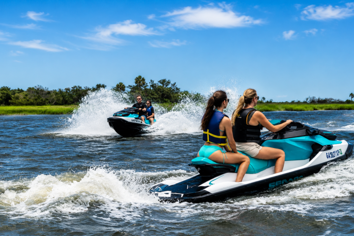 Two people on jet skis riding on a lake with blue skies and grassy shoreline.