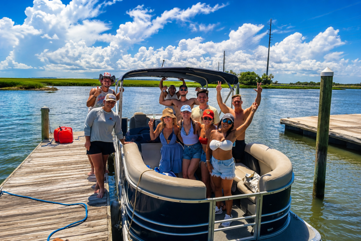 Group of people smiling on a pontoon boat at a dock, with blue sky and water.