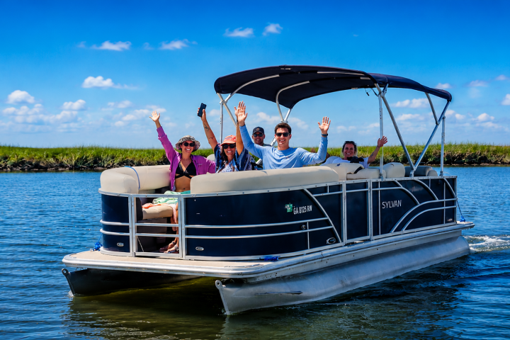 Group of people waving on a pontoon boat in calm water.