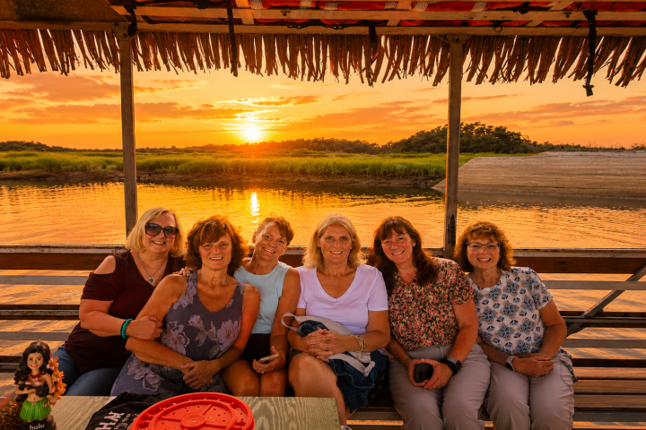 Group of six women sitting on a boat at sunset, smiling.
