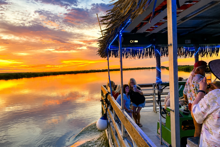 People on a boat with a straw roof enjoying a colorful sunset over calm water.