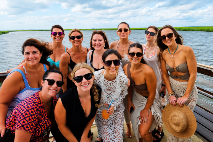 Group of smiling women in summer outfits on a boat near water.