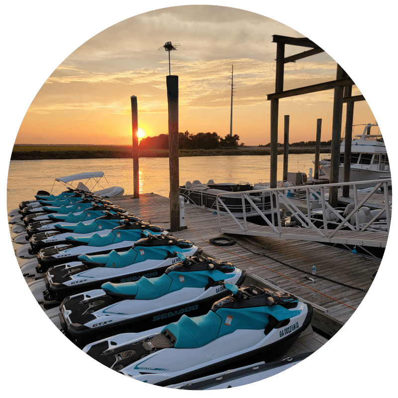 Rows of jet skis on a dock at sunset with boats in the background.