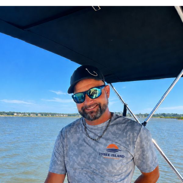 Person in cap and sunglasses on a boat with a sunny shoreline in the background.