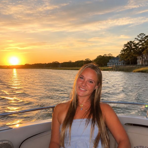 Woman smiling on a boat at sunset with calm water and trees in the background.