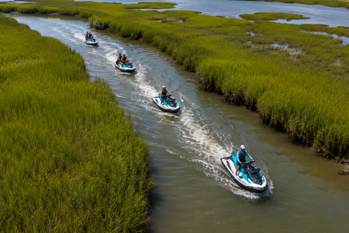 Four people ride jet skis in a narrow waterway surrounded by green marshland.